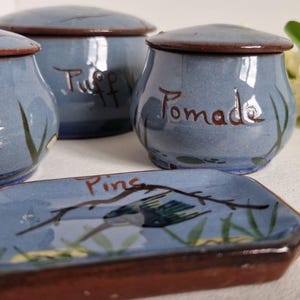 Puede incluir: Un conjunto de tarros de cerámica azules y una bandeja rectangular. Los tarros tienen tapas y la palabra "Pomade" está escrita en uno. La bandeja tiene un diseño de pájaro y rama. Todas las piezas tienen bordes y detalles marrones.