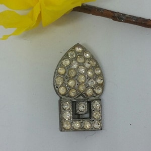 Puede incluir: Una hebilla de metal plateada con una parte superior en forma de corazón y una base rectangular. La hebilla está decorada con diamantes de imitación transparentes.