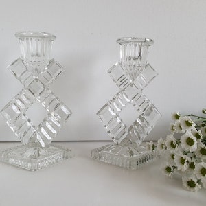 Könnte beinhalten: Zwei klare Glas-Kerzenhalter mit geometrischem Design. Die Halter sind aus facettiertem Glas und haben einen quadratischen Sockel.