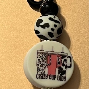May include: A black and white patterned keychain with a black carabiner clip, a black and white patterned bead, a white bead with the text "crazy cup lady" and a graphic of three cups, a black and white patterned bead, a black bead, and a silver key ring.
