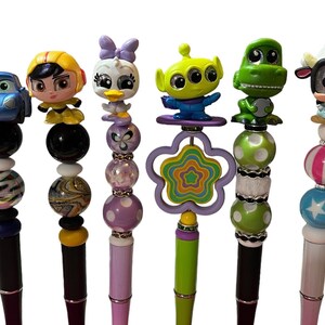 May include: Six colorful pens with different character toppers. The toppers include a blue car, a yellow female character, a purple duck, a green alien, a green dinosaur, and a black and white cow. The pens have various beads and designs.