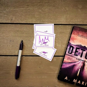 May include: A stack of white business cards with purple text and a feather design. The text on the cards reads "AM A. Marie Creative Works Co."