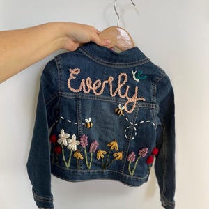 May include: A blue denim jacket with embroidered flowers, bees, and the name "Everly" in pink yarn.