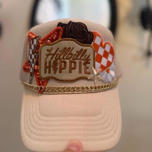 May include: A beige baseball cap with a curved brim, adorned with patches including a brown rose, a sequined orange shape, and a heart with an orange and white checkerboard pattern. The text "Hillbilly Hippie" is featured on a patch.