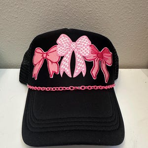 May include: Black trucker hat with a pink chain and three pink bows, one with a white checkered pattern.
