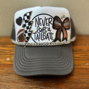 May include: A white and gray trucker hat with a black and white checkered lightning bolt, a brown bow with a football, a black heart, and the text "NEVER lost a TAILGATE" in a circle.