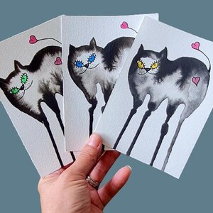 Puede incluir: Tres acuarelas de gatos negros con ojos de diferentes colores. Cada gato tiene un corazón rosa en la cola y está de pie sobre patas largas y delgadas.