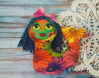 Puede incluir: Una sirena de peluche hecha a mano con un estampado floral colorido, cara verde y lazo rosa. La sirena tiene el pelo largo azul y una cola con estampado floral.