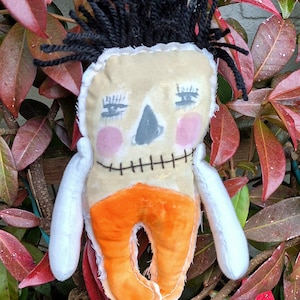 Könnte beinhalten: Eine handgemachte Stoffpuppe mit schwarzen Wollhaaren, einem gestickten Lächeln und einer orangefarbenen Stoffhose. Die Puppe ist mit ausgestreckten Armen posiert und sitzt in einem Bett aus roten und grünen Blättern.