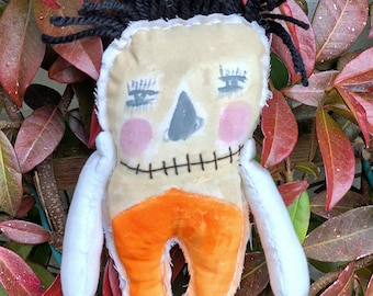 May include: A handmade rag doll with black yarn hair, a stitched smile, and orange fabric pants. The doll is posed with its arms outstretched and is sitting in a bed of red and green leaves.