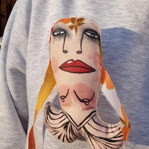 May include: A handmade fabric doll with a painted face, featuring red lips, dark eyes, and a floral headpiece. The doll has a unique design with a long, flowing orange and white fabric. The doll is attached to a gray sweatshirt.