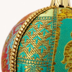 May include: A spherical Christmas ornament covered in multi-colored fabric featuring a woven pattern of teal, gold, and red. The surface is divided by a decorative gold braided trim that runs vertically along the side of the round ornament.