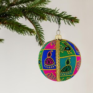 May include: A spherical Christmas ornament featuring a vibrant patchwork design of green, blue, and pink fabric squares. Each square displays embroidered paisley and peacock motifs in contrasting colors, accented by a gold metallic trim and cap.