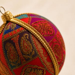 May include: A spherical Christmas ornament covered in fabric featuring a vibrant patchwork of red, purple, and blue panels with gold paisley and floral embroidery. A gold-toned metal cap and a textured gold ribbon trim accent the ornament.