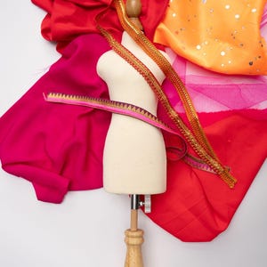 May include: A white dress form with a wooden base and a red, pink, and orange fabric draped over it. There are also gold and pink trims on the fabric.