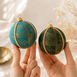May include: Two spherical ornaments featuring a grid pattern with intricate geometric and floral motifs. One ornament is teal and the other is dark green, both accented with gold-colored beaded vertical trim and a gold-colored metal cap.