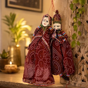 May include: Two Indian puppets, a male and female, hang from red strings. They wear maroon dresses with white dot patterns and colourful trim. The puppets have painted faces and are displayed against a wooden background.