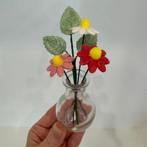 May include: A small glass vase with a round body holds a bouquet of handmade clay flowers. The flowers are pink, red, and white with yellow centres. The leaves are green.