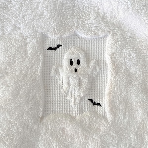 May include: White cross-stitch embroidery of a ghost with two black bats on a white background.