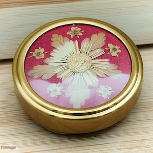 Peut inclure: Une boîte à bijoux ronde de couleur dorée avec un couvercle en verre. La boîte est décorée de fleurs séchées sur un fond rouge. Les fleurs sont blanches et brunes.