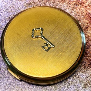 Könnte beinhalten: Ein runder, goldfarbener Kompaktspiegel mit einer texturierten, kreuzschraffierten Oberfläche. In der Mitte des Deckels befindet sich ein kleines, metallisch goldenes Schlüsselemblem, das die Zahl 21 im Design integriert.