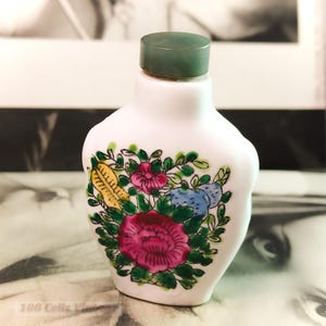 Op de afbeelding: Een kleine, witte keramische fles met een groene dop. De fles is versierd met een kleurrijk bloemmotief met roze, rode, gele en blauwe bloemen met groene bladeren. De fles is waarschijnlijk een decoratief item.