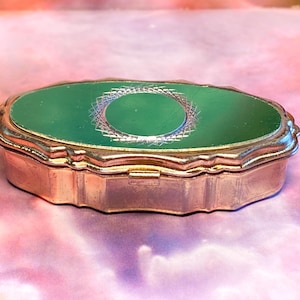 May include: An oval-shaped metal trinket box with a polished gold-tone finish and a scalloped edge. The lid features a smooth, deep green inlay centred with a circular, etched geometric pattern that radiates outward in a fine, cross-hatched design.