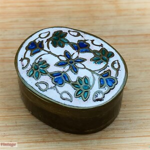 Op de afbeelding: Een klein, ovaalvormig messing sieradendoosje met een wit emaille deksel. Het deksel is versierd met een bloemmotief in blauw, groen en wit.