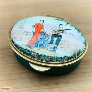 Puede incluir: Una caja de alhajas ovalada de color verde con detalles dorados. La tapa presenta una escena pintada de una mujer con un vestido rojo y azul bailando con un hombre que toca la guitarra. El fondo es un cielo azul claro con un edificio blanco y árboles verdes.