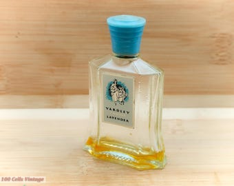 Puede incluir: Un frasco de perfume Yardley Lavender vintage. El frasco de vidrio transparente tiene una tapa azul claro y una etiqueta con una ilustración turquesa y blanca. El frasco contiene un líquido amarillo y está sobre una superficie de madera.