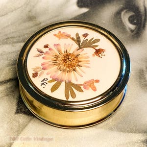 May include: A vintage round compact with a gold-tone base and a floral design on the lid. The lid features a pressed flower arrangement with pink and brown hues, set against a white background. The compact has a black rim and a small signature.