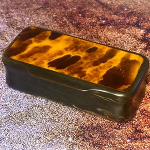May include: A rectangular box with a dark brown, smooth base and a lid featuring a mottled, amber and dark brown tortoiseshell-like pattern. The lid sits flush with the base, and the entire object has a polished, glossy finish throughout.
