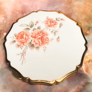 May include: A round, white compact mirror with a scalloped edge and a gold-tone metal rim. The top surface features a floral illustration of pink roses with green leaves and stems, arranged in a cluster on the left side of the white background.