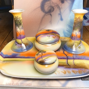 Könnte beinhalten: Ein Porzellan-Toilettenset bestehend aus zwei Kerzenhaltern, zwei Döschen mit Deckel und einem rechteckigen Tablett. Jedes Teil zeigt eine handgemalte Landschaft in Orange-, Gelb- und Blautönen mit Bäumen und Wasser bei Sonnenuntergang.