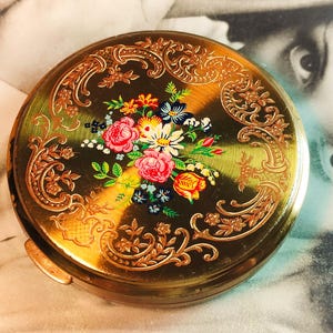 Puede incluir: Un estuche compacto dorado vintage con un diseño floral. El estuche compacto presenta un ramo central de flores rosas, rojas, amarillas y azules. Los bordes de filigrana dorada adornan el arreglo floral y el borde del estuche.