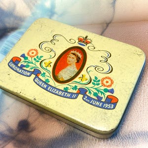 Op de afbeelding: Een rechthoekig, crèmekleurig metalen blik met een centraal ovaal portret van koningin Elizabeth II. Het deksel toont een kroon, bloemmotieven en een blauwe lintbanner met de tekst CORONATION QUEEN ELIZABETH II 2ND JUNE 1953.