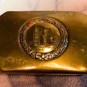 May include: A rectangular brass box with beveled corners and a polished finish. The lid features a central circular medallion depicting an architectural structure, surrounded by a detailed relief of a laurel wreath tied with a bow at the bottom.