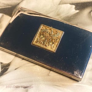 Puede incluir: Un pitillera rectangular plateada con una superficie azul oscuro. Un cuadrado dorado con un diseño floral ornamentado está centrado en la pitillera. La pitillera es ligeramente reflectante, mostrando signos de edad.