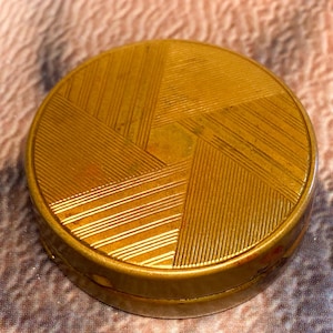 Peut inclure: Un poudrier rond en métal doré avec un motif géométrique de lignes parallèles gravées sur le couvercle. Le design se compose de sections triangulaires disposées en forme de moulinet, créant une surface texturée sur le dessus.