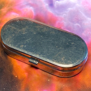 May include: A small, oval-shaped metal container with a hinged lid. The exterior surface of the silver-toned box shows visible signs of wear, including fine scratches and minor surface imperfections across the top and along the side edges.