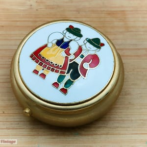 May include: A gold-coloured, round trinket box with a white lid featuring a colourful enamel design of two figures in traditional folk attire. The figures are dancing. The box is on a wooden surface.