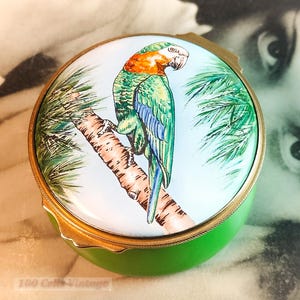 May include: A circular, green trinket box with a lid depicting a hand-painted parrot on a branch. The parrot is painted in green, blue, and orange. The box has a gold-coloured rim and the text "100 Cells Vintage".