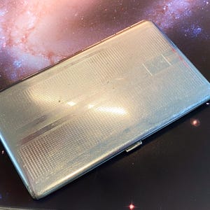 May include: A rectangular silver-toned metal cigarette case featuring a fine cross-hatched grid pattern across its surface. The case has a smooth, polished border and a small metal latch on the side for opening and closing the hinged container.