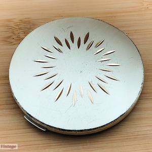 Pode incluir: Um estojo compacto de prata com um design circular que apresenta um padrão de raios de sol. O estojo tem um acabamento liso e metálico.