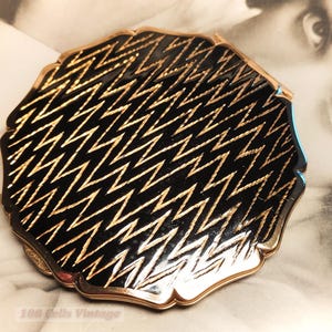 May include: Vintage compact with a black and gold zigzag pattern. The compact has a scalloped edge and a gold-toned frame. The design is a repeating pattern of gold zigzags on a black background. The compact is a classic vintage accessory.