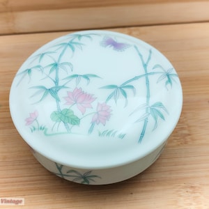 Puede incluir: Una caja redonda de cerámica blanca con tapa. La tapa presenta un diseño pintado de tallos de bambú, flores de loto rosas y una mariposa morada. La caja está sobre una superficie de madera. El texto "100 Cells Vintage" está en la parte inferior.