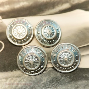 Puede incluir: Cuatro botones plateados con un diseño de rueda y la inscripción "BRITISH RAILWAYS" en el borde. Los botones circulares tienen un aspecto vintage y ligeramente deslustrado, evocando el pasado.