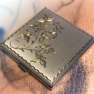 Pode incluir: Um estojo compacto quadrado de metal prateado com acabamento escovado e um padrão floral gravado em tom dourado na tampa. As bordas da tampa são contornadas por uma linha gravada festonada e um pequeno fecho de metal é visível.