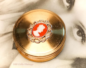 May include: A vintage gold-coloured compact with a cameo design. The compact features a central oval cameo with a white profile against a red background, framed by a decorative silver-coloured border. The compact is round and has a brushed metal finish.