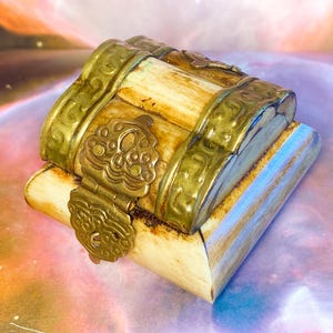 May include: A small, rectangular box with a curved, hinged lid, constructed from cream-coloured panels with visible grain. The box is reinforced with ornate, embossed brass metal bands and a central brass latch featuring intricate, decorative patterns.